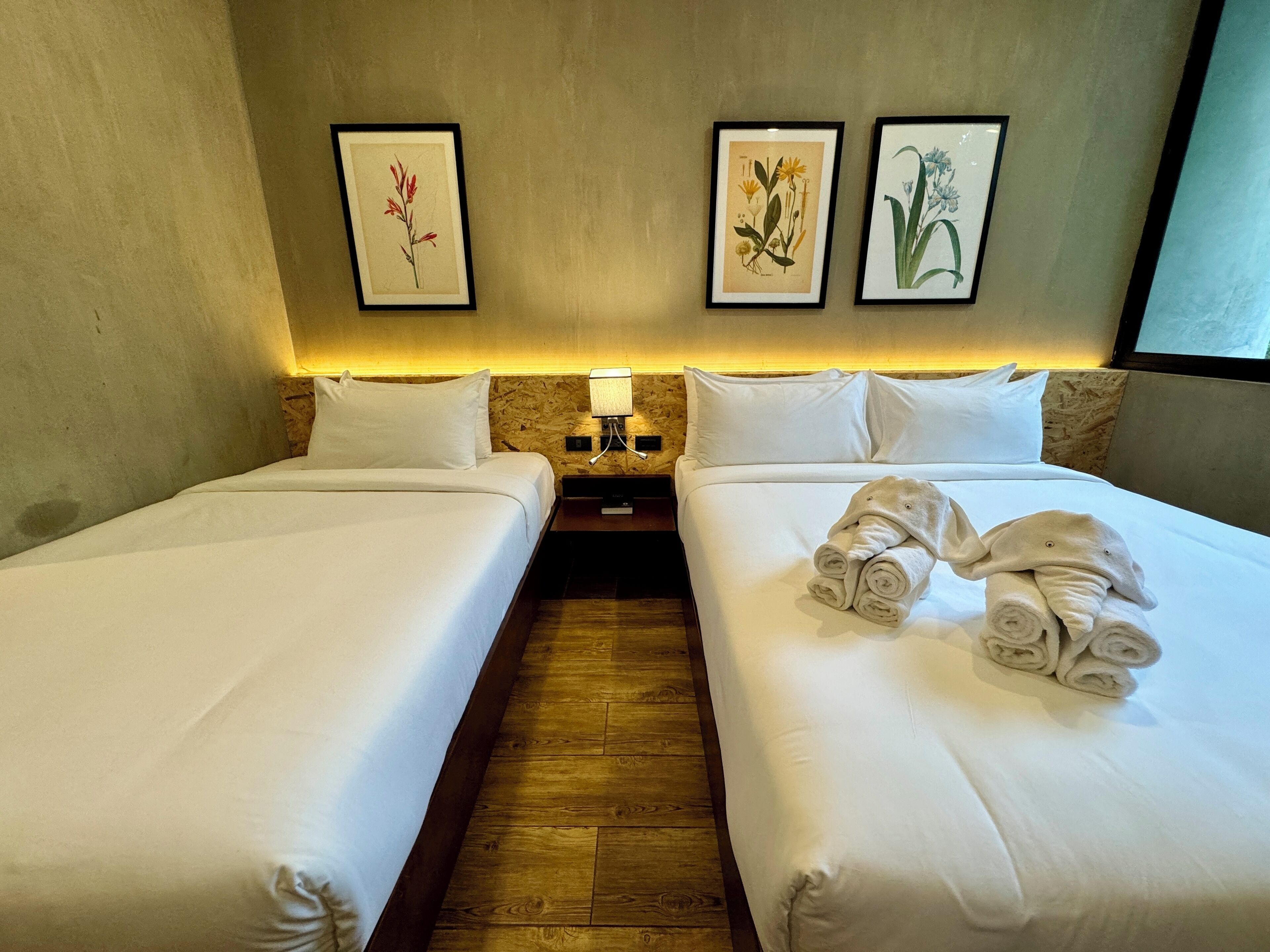 Ein Hotelzimmer mit zwei Betten, weißen Leinentüchern, Holzboden, einem Nachttisch mit Lampe und drei gerahmten Pflanzenbildern an der Wand.