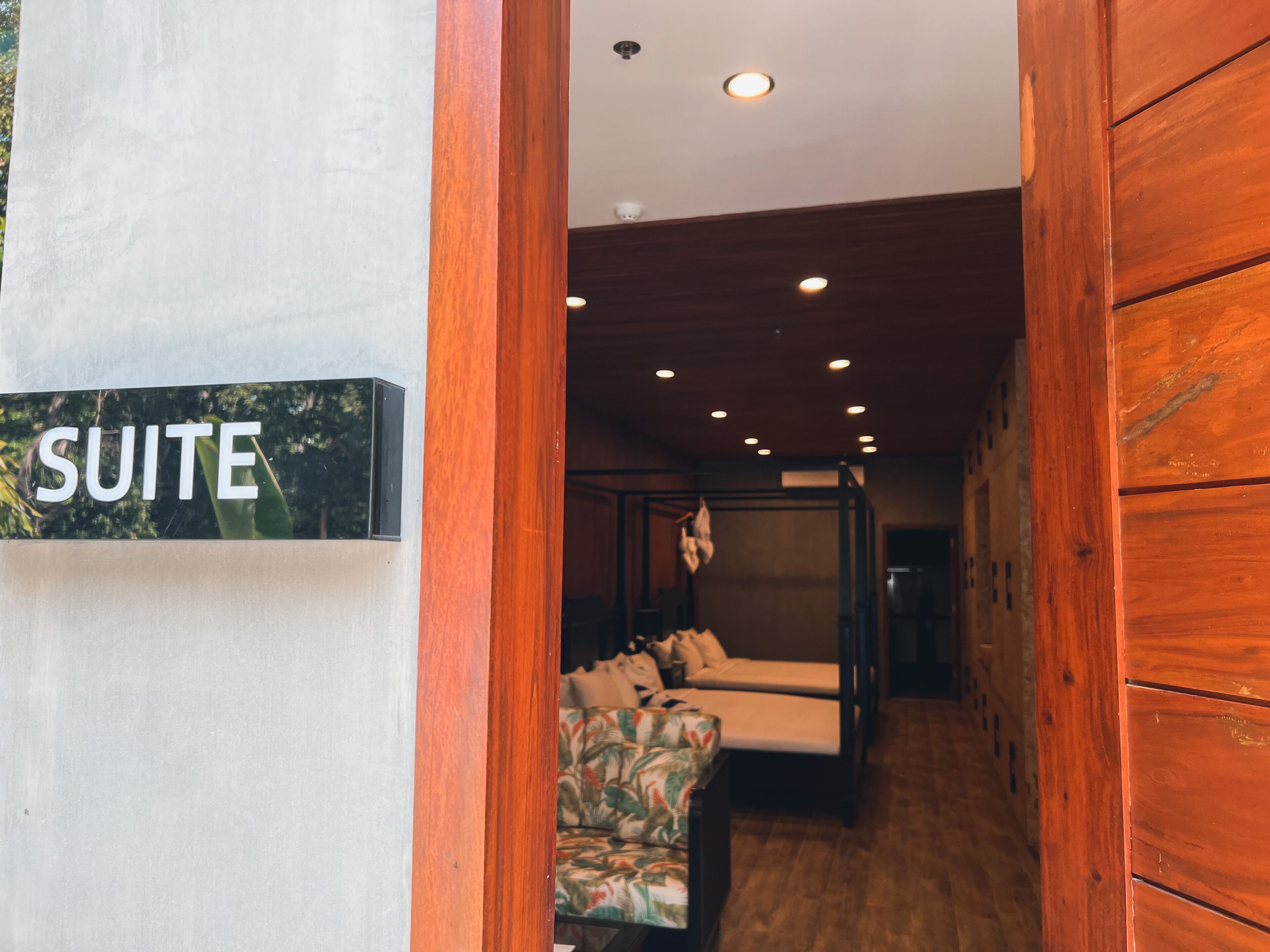 A wooden-framed room with a bed and a sofa, a sign reading "SUITE", and a ceiling with recessed lighting.