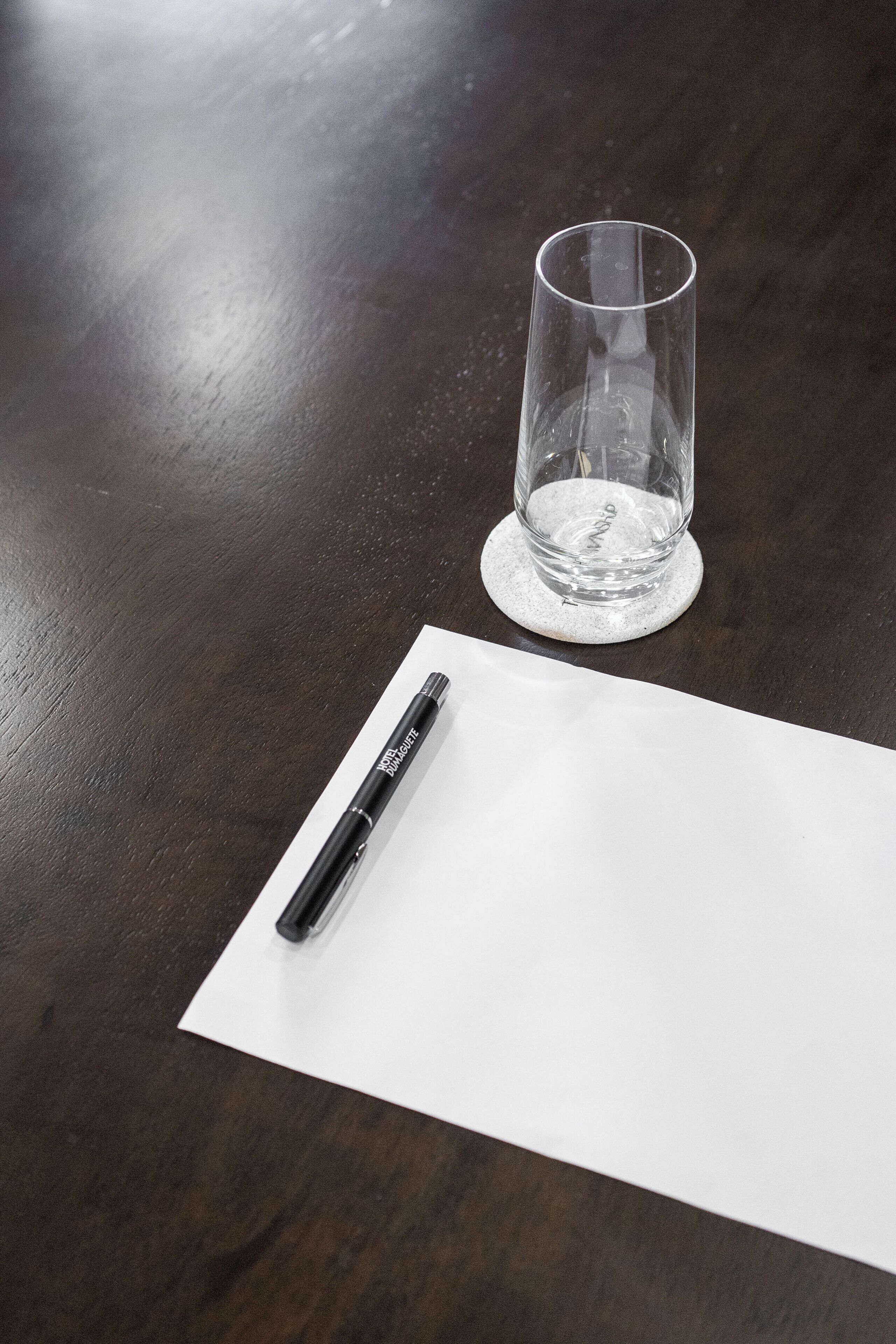A clear glass on a coaster, a pen on a white paper, and a dark wooden table.