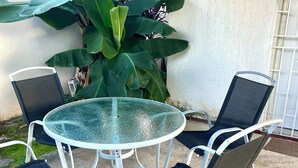 Une table ronde en verre, entourée de chaises en métal, est placée sur une terrasse en béton, avec une grande plante en arrière-plan.