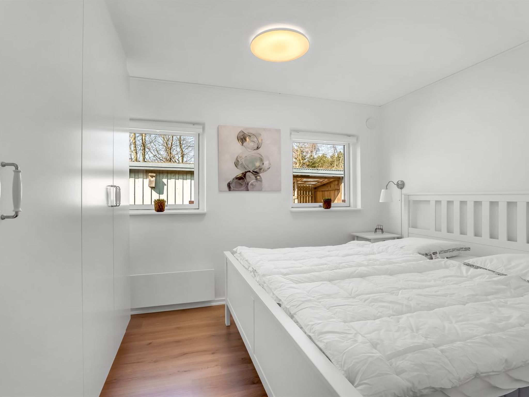 A bedroom with a large bed, two windows, a ceiling light, and a small bedside table.