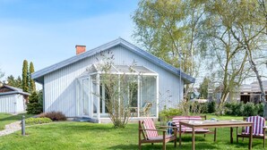 Een modern huis met een glazen conservatorium, een terras met houten meubels en een verzorgd gazon.