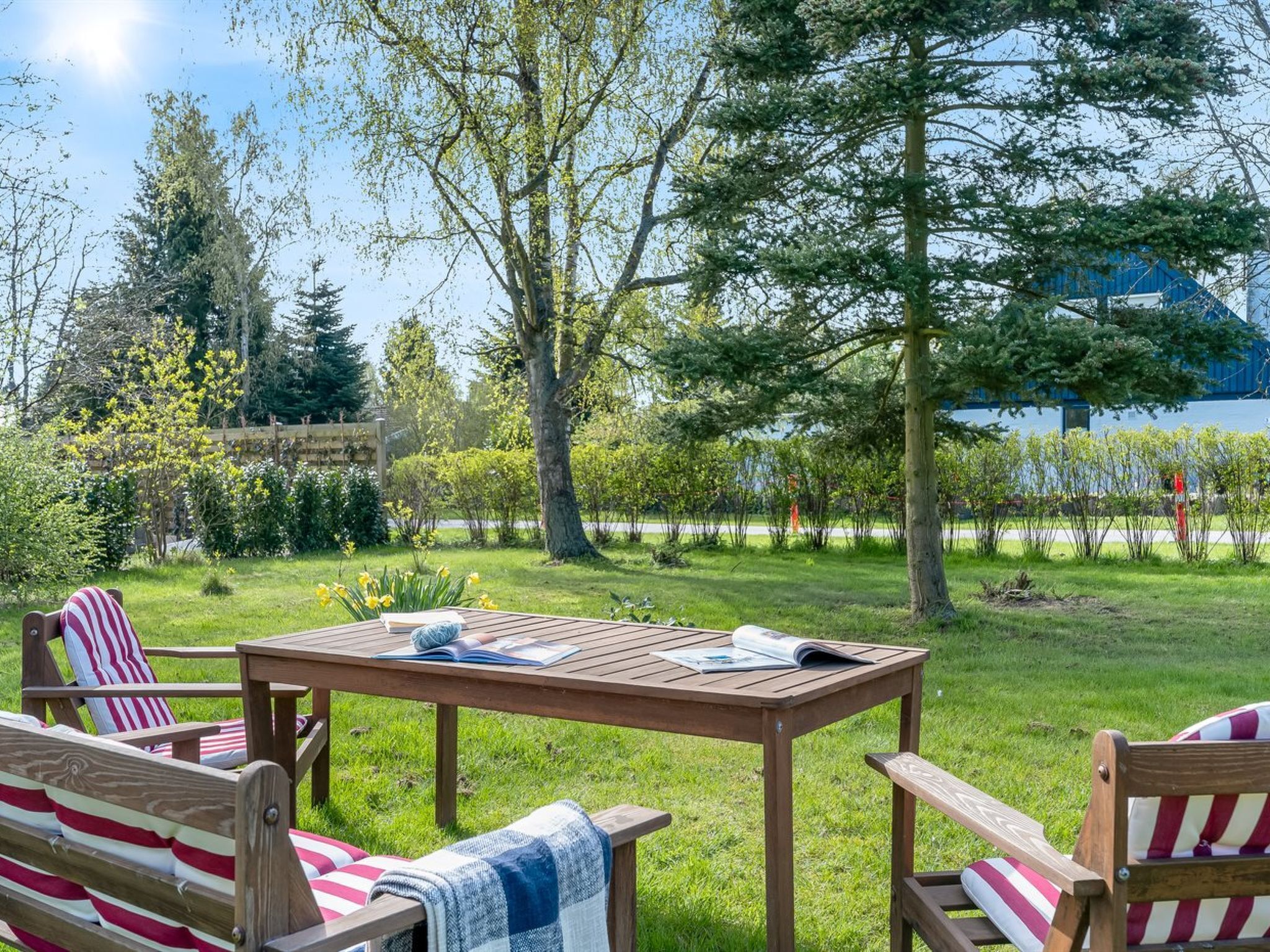 A wooden table with a book and magazine, surrounded by striped chairs in a grassy outdoor area with trees and a blue building in the background.