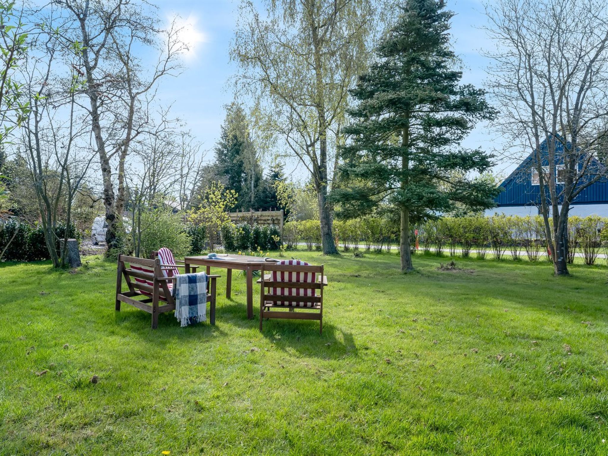 Een terras buiten met een houten tafel en bankjes, omringd door bomen en een gazon.