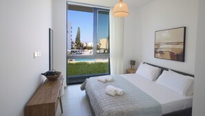 A modern hotel room with a large bed, a wooden side table, a view of a pool and buildings, and a hanging light fixture. - Protaras Aqua Pearl Villa AQ01 (Protaras)