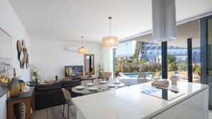 A modern kitchen with a central island, dining area, and a view of a pool and garden. - Protaras Aqua Pearl Villa AQ01 (Protaras)