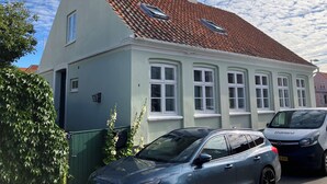 Une maison à deux étages, avec un toit recouvert de tuiles rouges, de nombreuses fenêtres et une voiture garée dans l’allée.