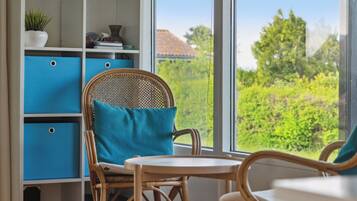 A room with a wicker chair, a small table, a bookshelf with blue storage boxes, and a view of greenery outside.