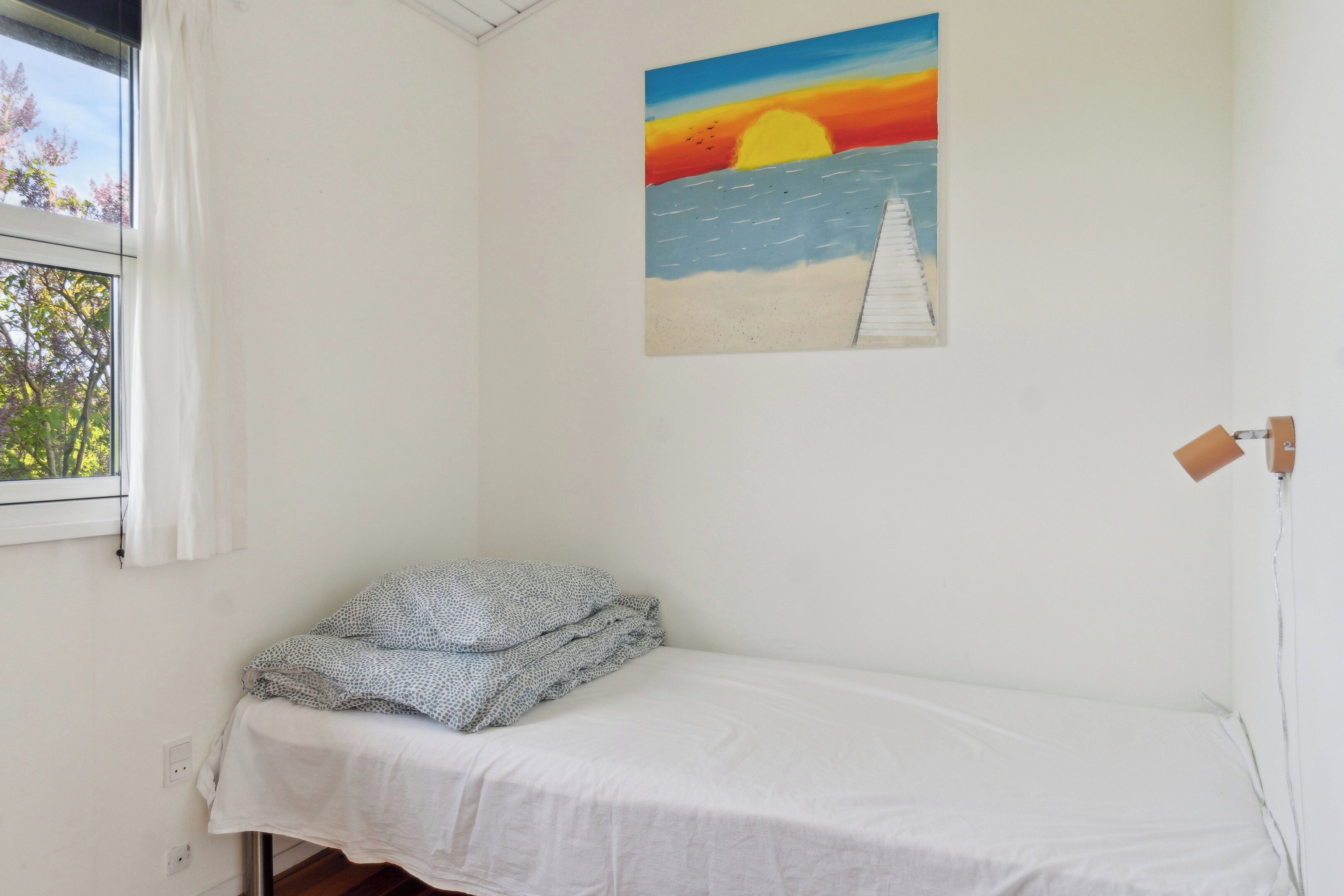 Uma cama de solteiro com lençol branco, um quadro de um pôr do sol sobre o oceano e uma janela com vista para árvores.