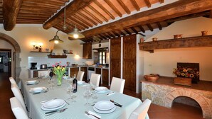 Zona pranzo con tovaglia a quadri, sedie bianche e un vaso di fiori. Sullo sfondo, una cucina con pensili in legno e un lampadario.