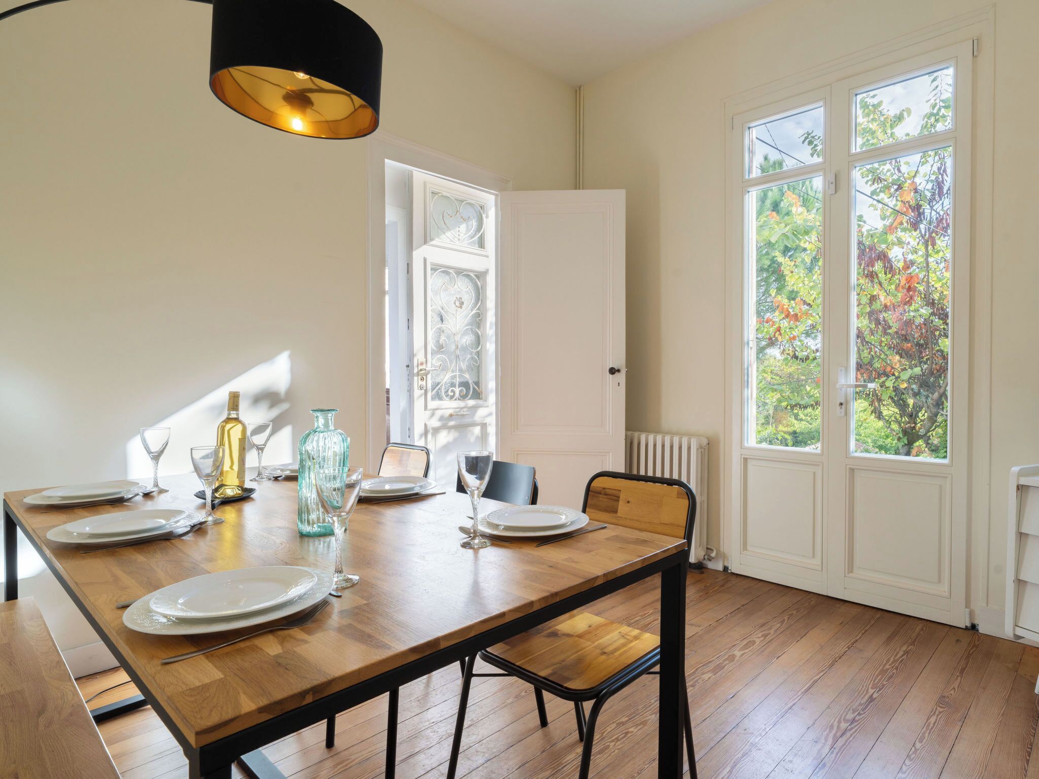 A dining area with a wooden table set for four, a black pendant light, and a view of a garden through a large window.