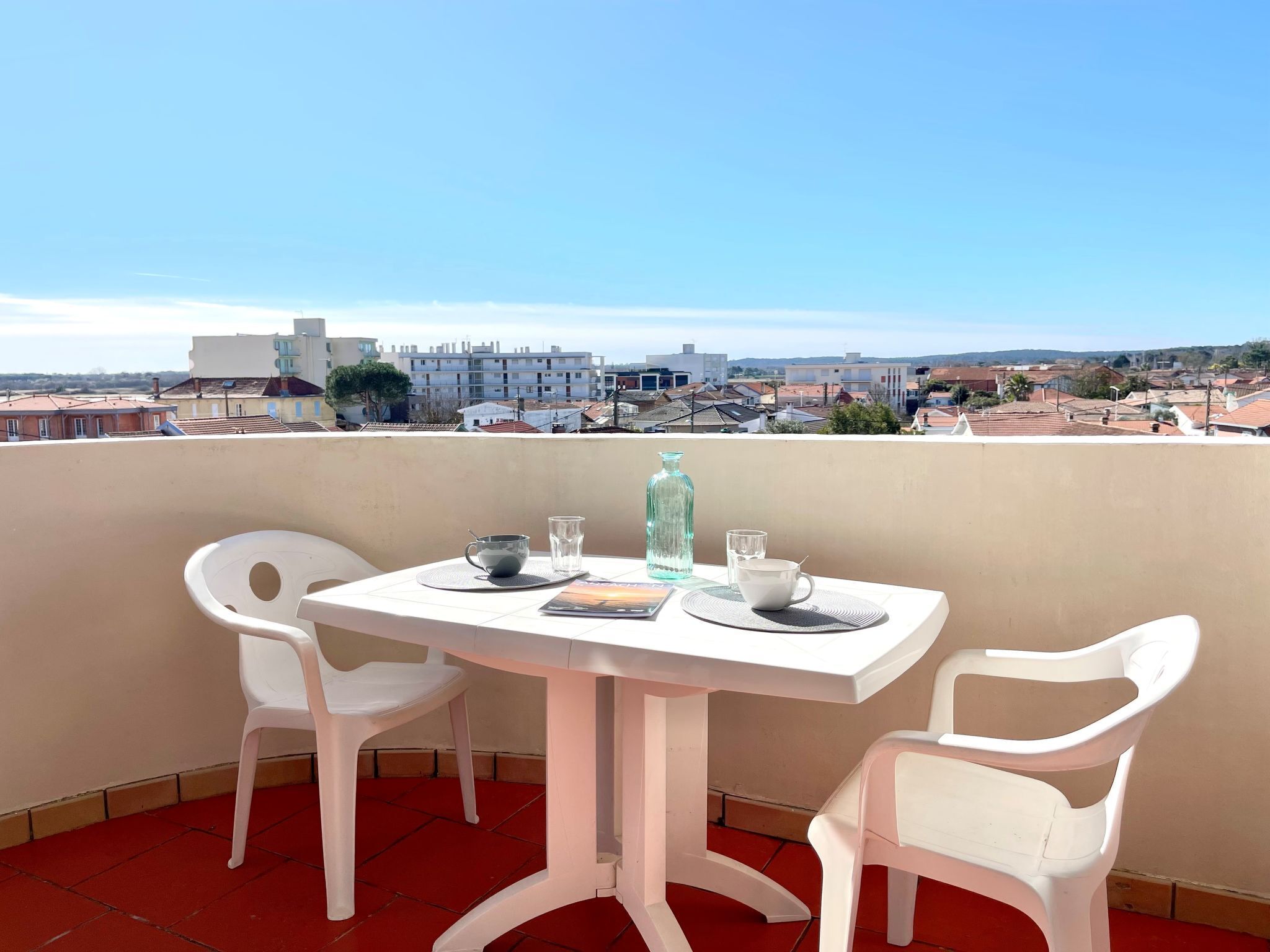 Un balcon avec une table et deux chaises blanches, donnant sur un quartier résidentiel avec des immeubles et un ciel dégagé.