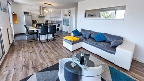 Un salon moderne avec un canapé d’angle, une table basse en verre et un coin repas, la cuisine étant visible au fond.