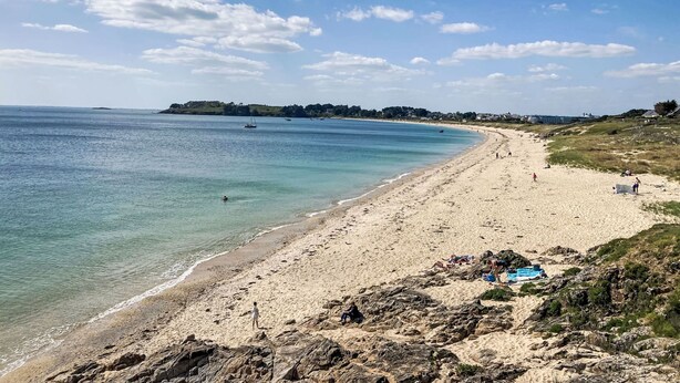 Une plage aux eaux cristallines, un rivage sablonneux et une côte lointaine parsemée de bâtiments.