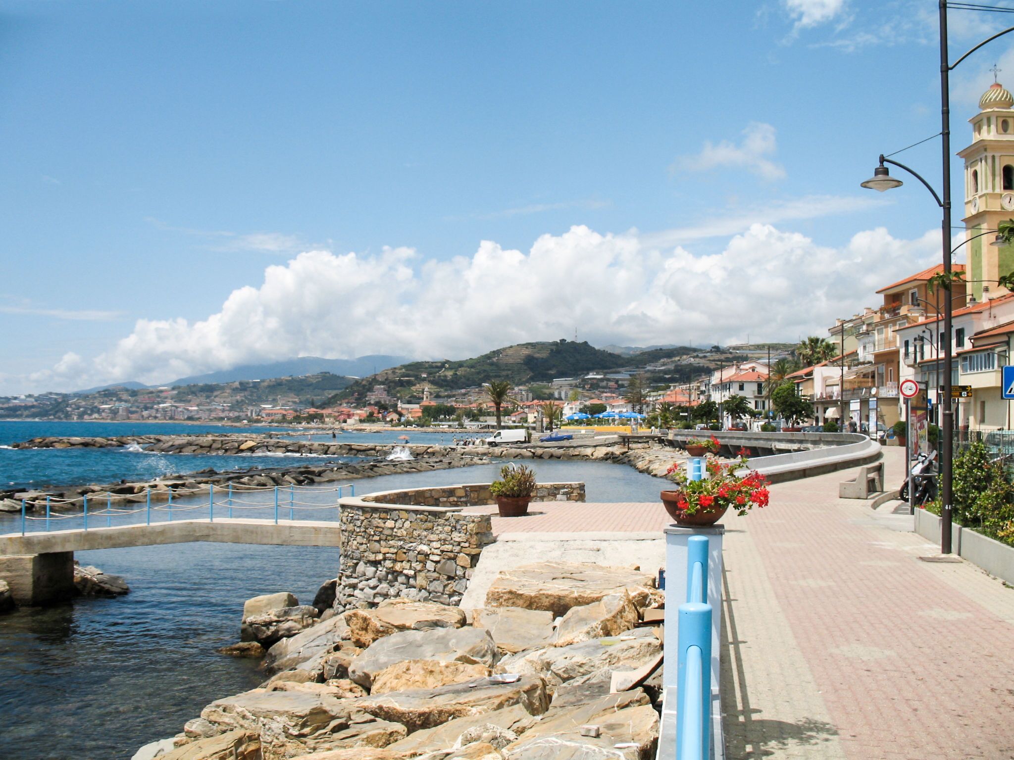 A coastal town with a stone bridge over a rocky harbor, a paved walkway, and buildings with Mediterranean architecture.