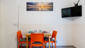 Un coin repas avec une table dressée pour quatre personnes, des chaises orange, un tableau accroché au-dessus de la table et un téléviseur fixé au mur.