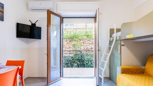 Une petite pièce comprenant un lit, un bureau et une chaise. Il y a une fenêtre donnant sur un jardin et un mur en pierre.