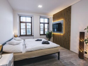Ein modernes Schlafzimmer mit einem Bett, zwei Fenstern, einem an einer Holzplatte montierten Fernseher und einer Pflanze.