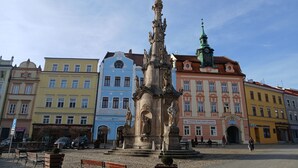 A public square with a historical monument, colorful buildings, and a clock tower.
