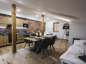 A modern kitchen with exposed brick walls and wooden beams, a dining area with a wooden table and chairs, and a bed with a white headboard.