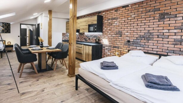 A modern room with a brick wall, a wooden dining table with chairs, a bed with towels, and a kitchen area.