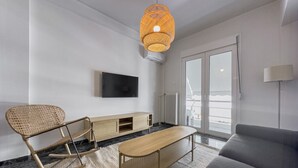 A modern living room with a sofa, a wooden coffee table, a wicker rocking chair, a TV mounted on the wall, and a large window with a view. - Phaedrus Living: Koukaki Verdant Marble (Athina)