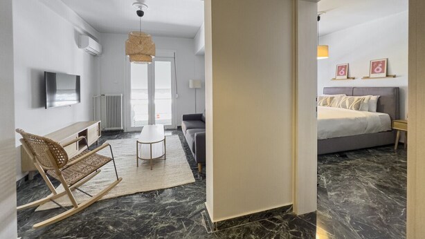 A hotel room with a bed, a sofa, a TV, a dining table, and a wicker chair. - Phaedrus Living: Koukaki Verdant Marble (Athina)