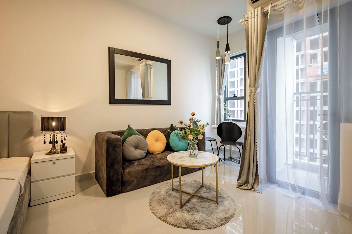 Un salon moderne comprenant un canapé, une table basse ronde, une table d’appoint avec une lampe, un miroir et une grande fenêtre avec des rideaux.