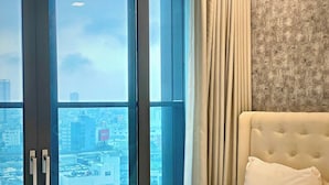 Habitación de hotel con un ventanal grande, una cama con cabecero blanco y vistas a un paisaje urbano.