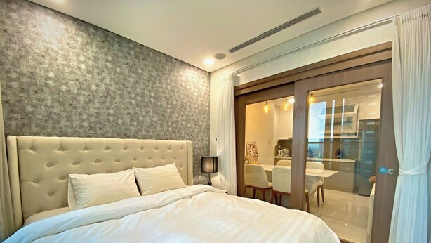 Un dormitorio con tablero de cama capitonado, una cama grande, una mesita de noche con lámpara y una puerta corrediza de vidrio que da a un comedor.