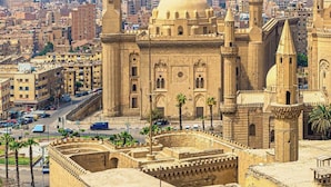 Een historisch gebouw met een grote koepel en meerdere minaretten, omgeven door een versterkte muur en palmbomen, met een piramide op de achtergrond.