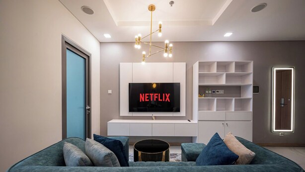 A modern living room with a large flat-screen TV, a green sofa, and a minimalist shelving unit. - DE LA SOL Centre Point By An Gia Hospitality (Ho Chi Minh City)
