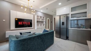 A modern living room with a large flat-screen TV, a blue sofa, and a kitchen area with a refrigerator and microwave. - DE LA SOL Centre Point By An Gia Hospitality (Ho Chi Minh City)