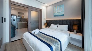 A modern hotel room with a large bed, a nightstand with a lamp, a painting on the wall, and a view of the kitchen and living area through an open door. - DE LA SOL Centre Point By An Gia Hospitality (Ho Chi Minh City)