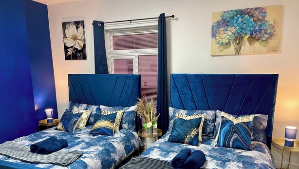A bedroom with two beds, each with a blue headboard and matching bedding. There are two bedside tables with lamps, a window with blinds, and artwork on the walls. - Modern 3 Bed Home ,Families and Contractor Stay with Free Parking (Kingston upon Hull)