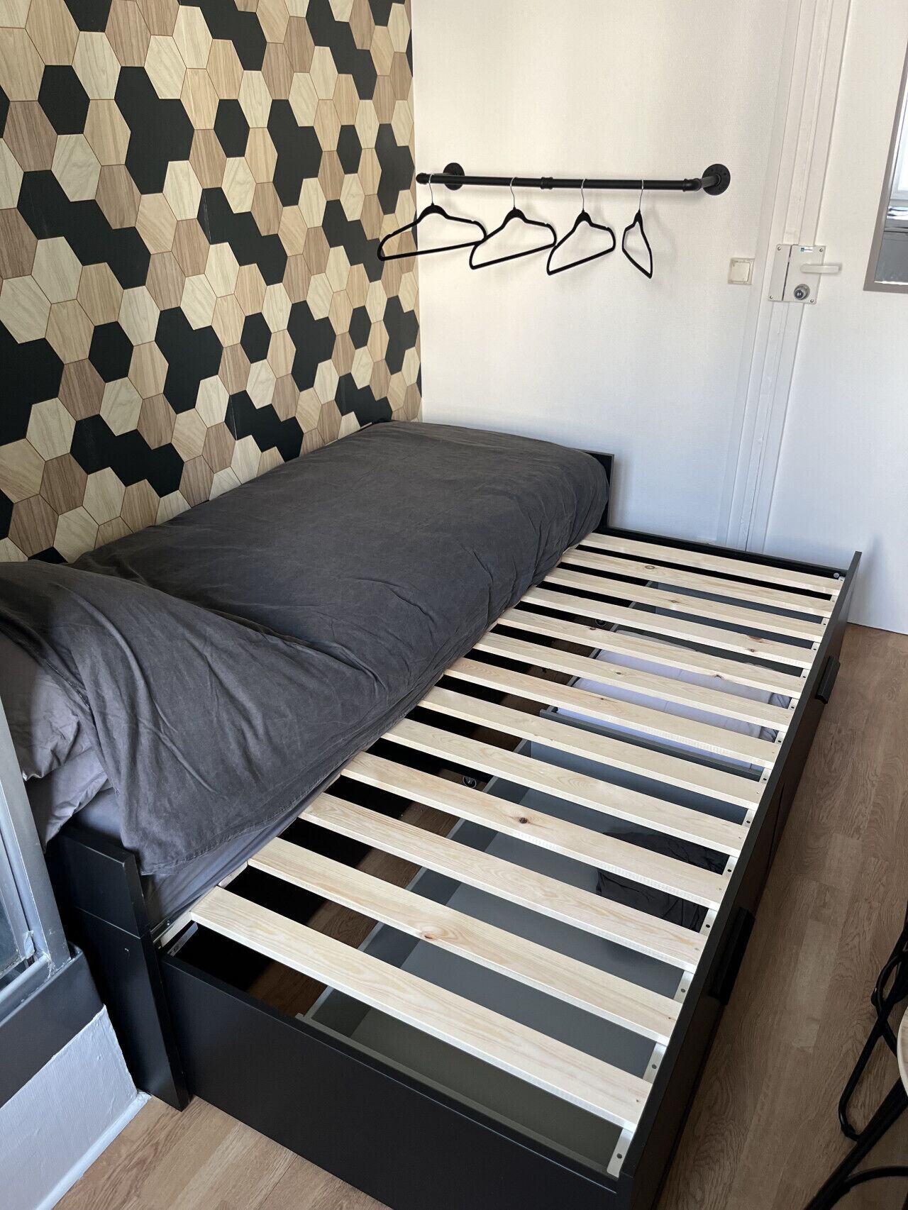 Un lit compact avec un cadre en bois et un matelas gris, un mur orné d’un motif géométrique et une porte dotée d’une poignée.