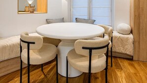 Een eetruimte met een ronde tafel en vier stoelen, een groot raam met zonwering, een houten vloer en een spiegel met wandlampen.