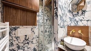 Een badkamer met een marmeren spatwand, houten kastjes, een glazen douchecabine en een witte wastafel met gouden details.