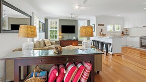 Una sala moderna con una mesa de centro de vidrio, un sofá y un televisor. Cuenta con una zona de cocina con armarios blancos y un comedor con taburetes.
