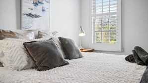 Una cama bien hecha con un edredón blanco estampado, dos almohadas y una manta enrollada a un lado. Encima de la cama, en la pared, hay un cuadro con una escena costera.