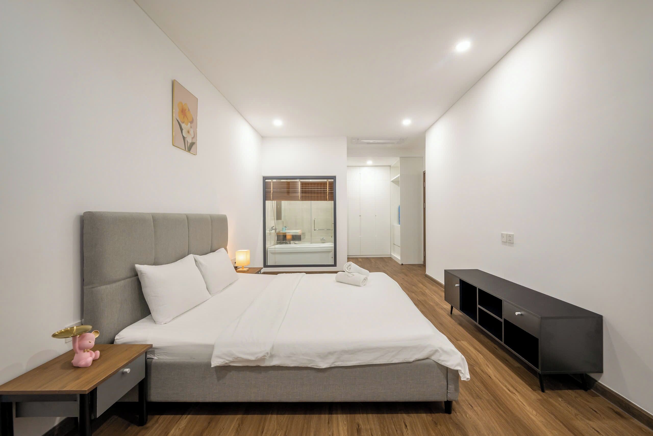 A modern bedroom with a grey bed, white bedding, a bedside table with a lamp, and a black TV stand.