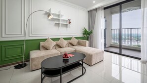 A modern living room with a beige sectional sofa, a black coffee table, and a floor lamp. There is a large window with a view of a cityscape and a balcony with a railing. - OPAL SAIGON PEARL Premier Residence (Ho Chi Minh City)