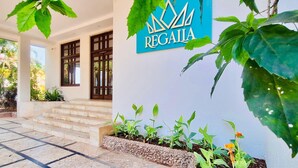 A building entrance with a sign reading "REGAIA," steps leading up to the entrance, and a garden area with plants and a small flower. - The Regalia Goa (Alto de Porvorim)