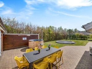Un jardin avec une terrasse en bois, un jacuzzi, un trampoline et une table entourée de chaises jaunes.