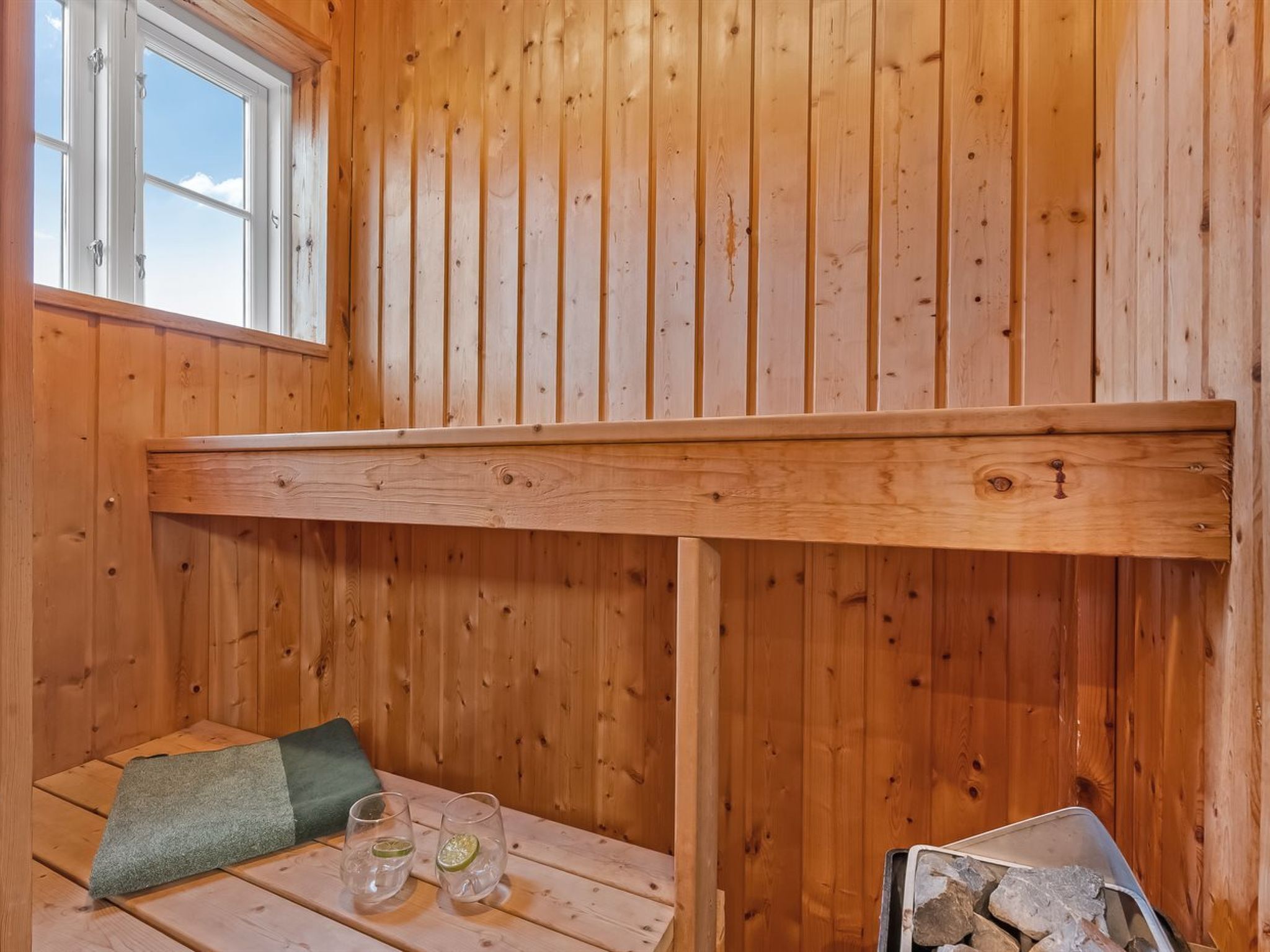 A wooden cabin interior with a loft bed, a window, a table with glasses and a green cushion.