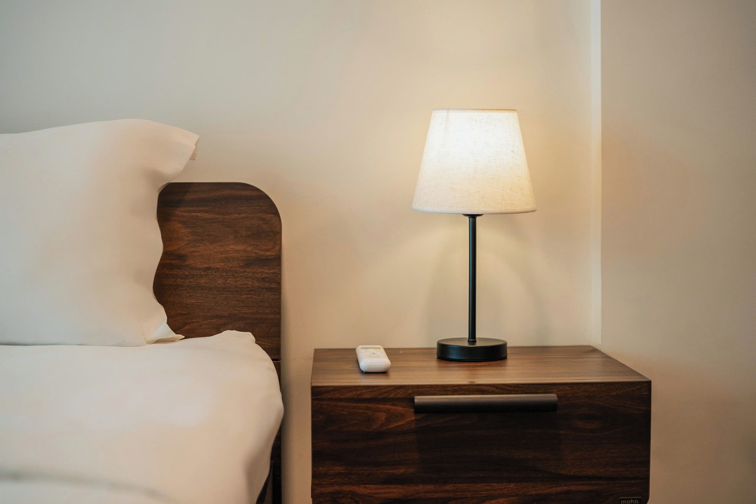 A neatly made bed with white linens, a wooden nightstand with a lamp, and a remote control on the nightstand.
