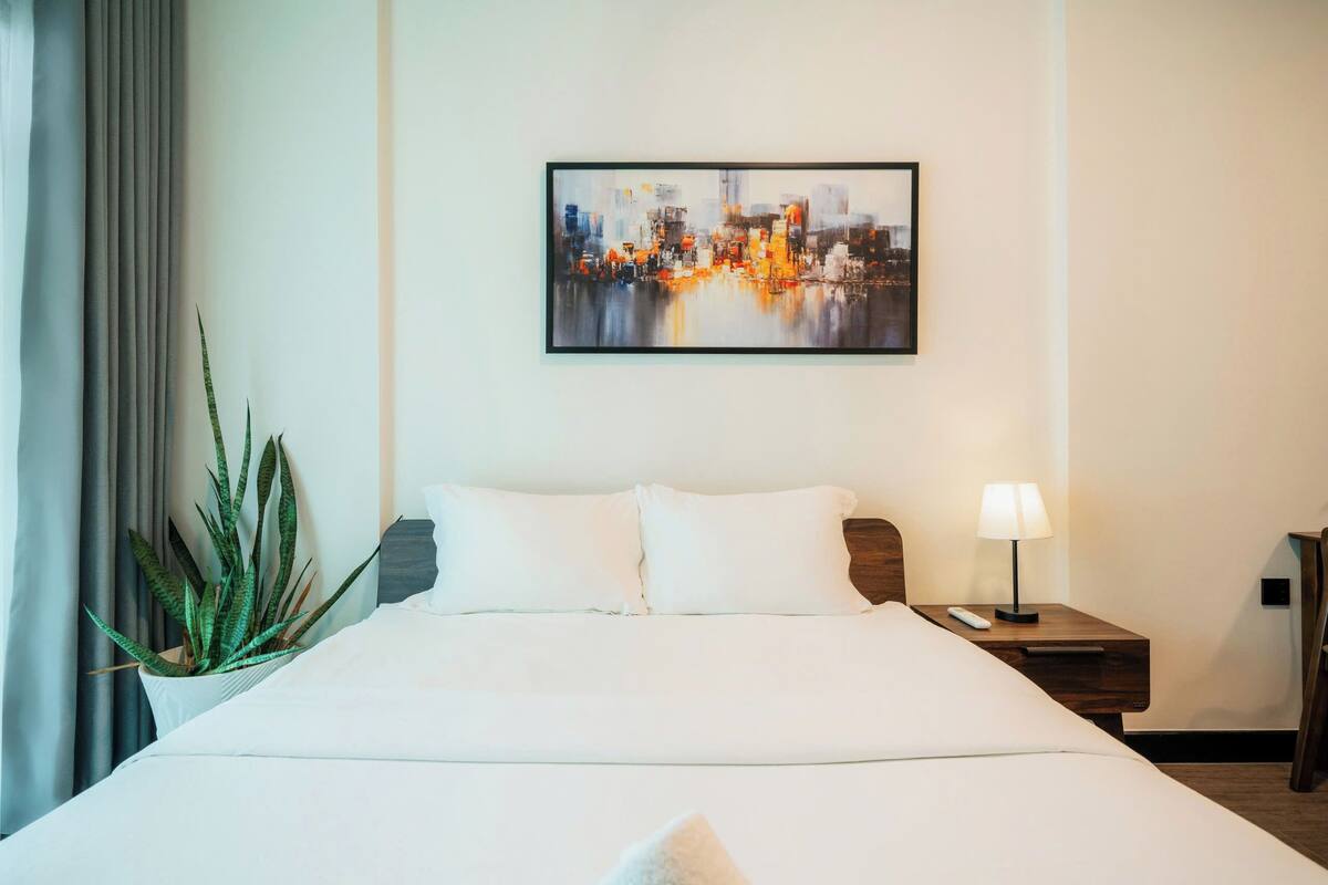 Una camera d'hotel con un letto grande, un comodino con una lampada, un telecomando e un quadro di un panorama cittadino appeso al muro.