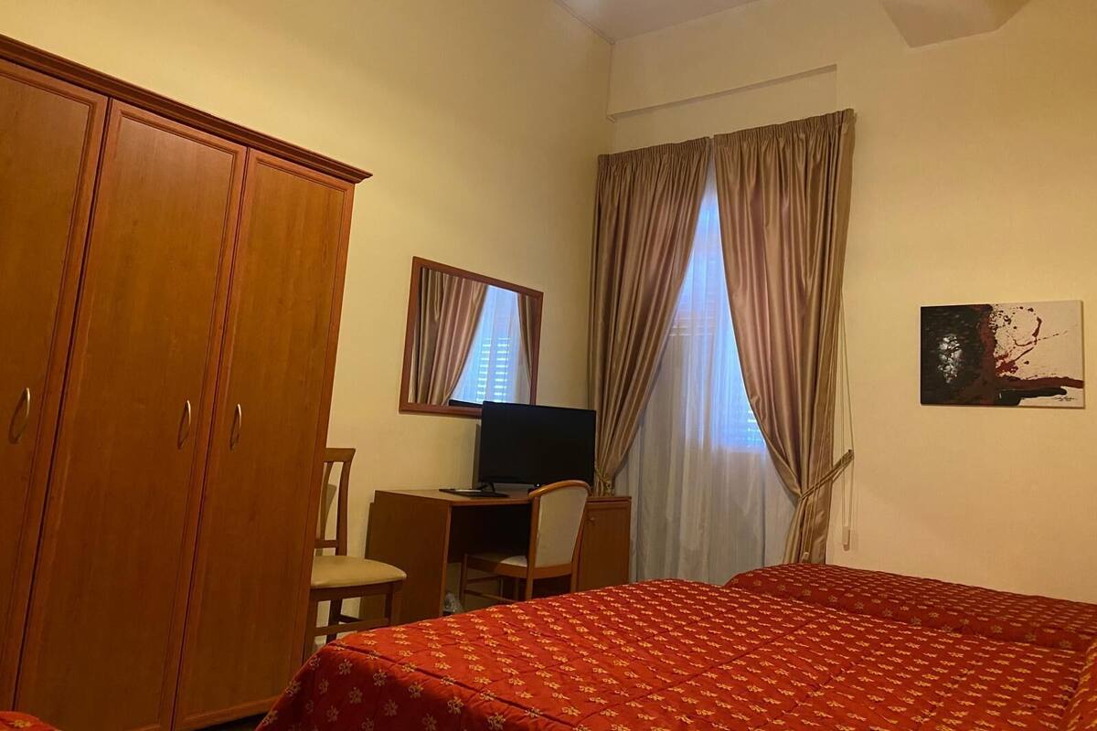 Una habitación de hotel con cama, escritorio, silla, armario, ventana con cortinas y un cuadro en la pared.