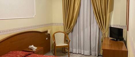Una habitación de hotel con cama, una silla, un escritorio y televisor, además de cortinas.