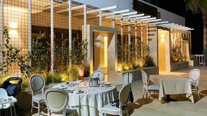 An outdoor dining area with tables and chairs, surrounded by a modern building with a tiled facade and warm lighting.